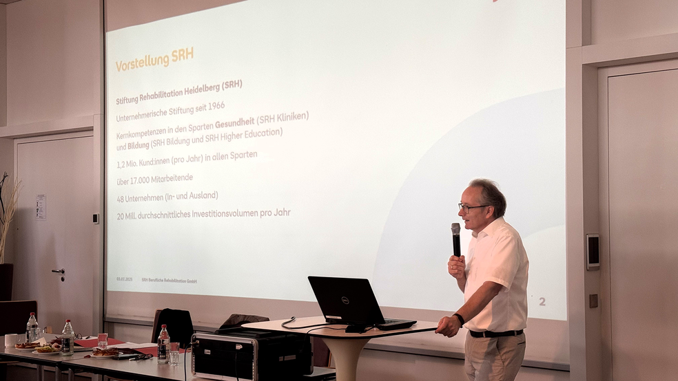 Dr. Gustav Wirtz hält eine Präsentation mit einem Mikrofon in der Hand, während eine Folie mit Informationen hinter ihm angezeigt wird.