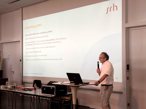 Dr. Gustav Wirtz hält eine Präsentation mit einem Mikrofon in der Hand, während eine Folie mit Informationen hinter ihm angezeigt wird.