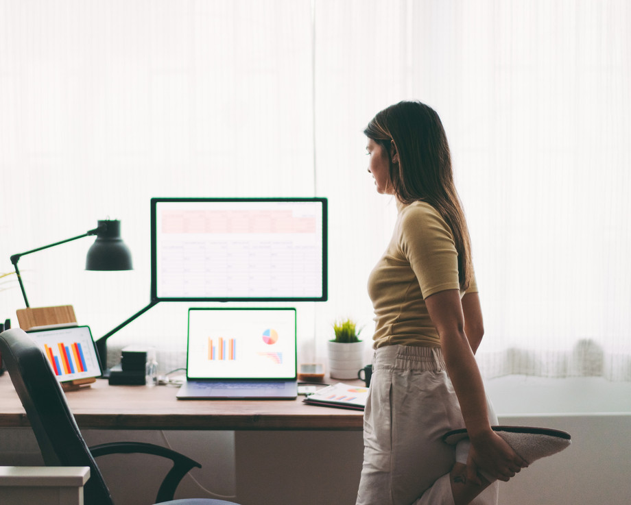 Frau steht an einem Schreibtisch mit Computer und Laptop.